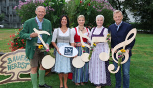 Fünf Personen in traditioneller Tracht halten Noten und Blumen in den Händen und stehen auf einer Wiese neben einem Schild mit der Aufschrift „Bauernherbst“.