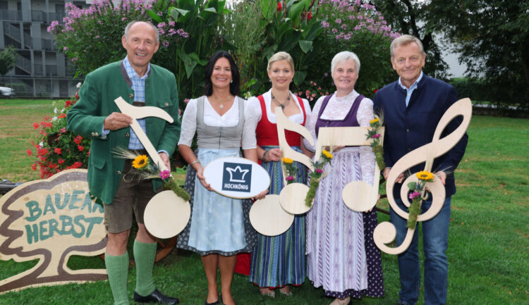 Fünf Personen in traditioneller Tracht halten Noten und Blumen in den Händen und stehen auf einer Wiese neben einem Schild mit der Aufschrift „Bauernherbst“.