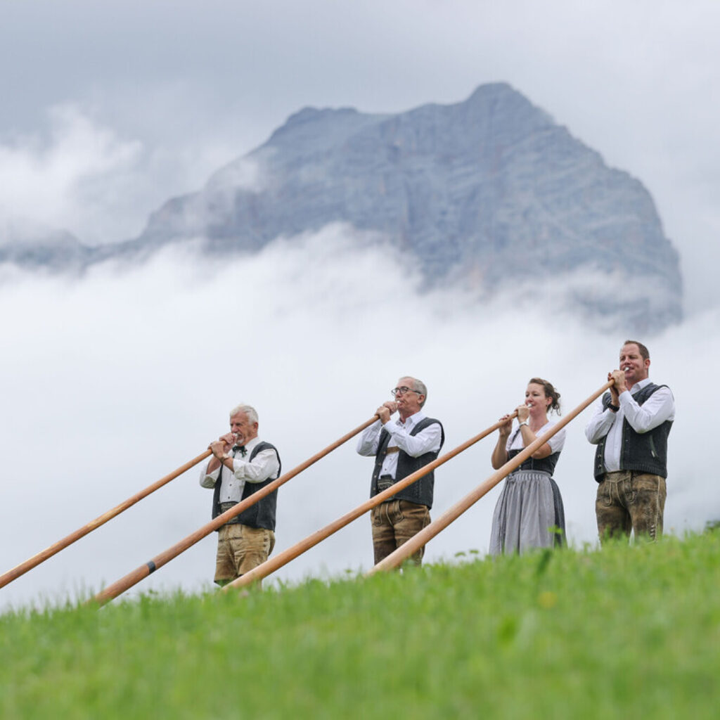 Vier Menschen spielen Alphorn auf einem grasbewachsenen Hügel, im Hintergrund ist ein nebelverhüllter Berg zu sehen.