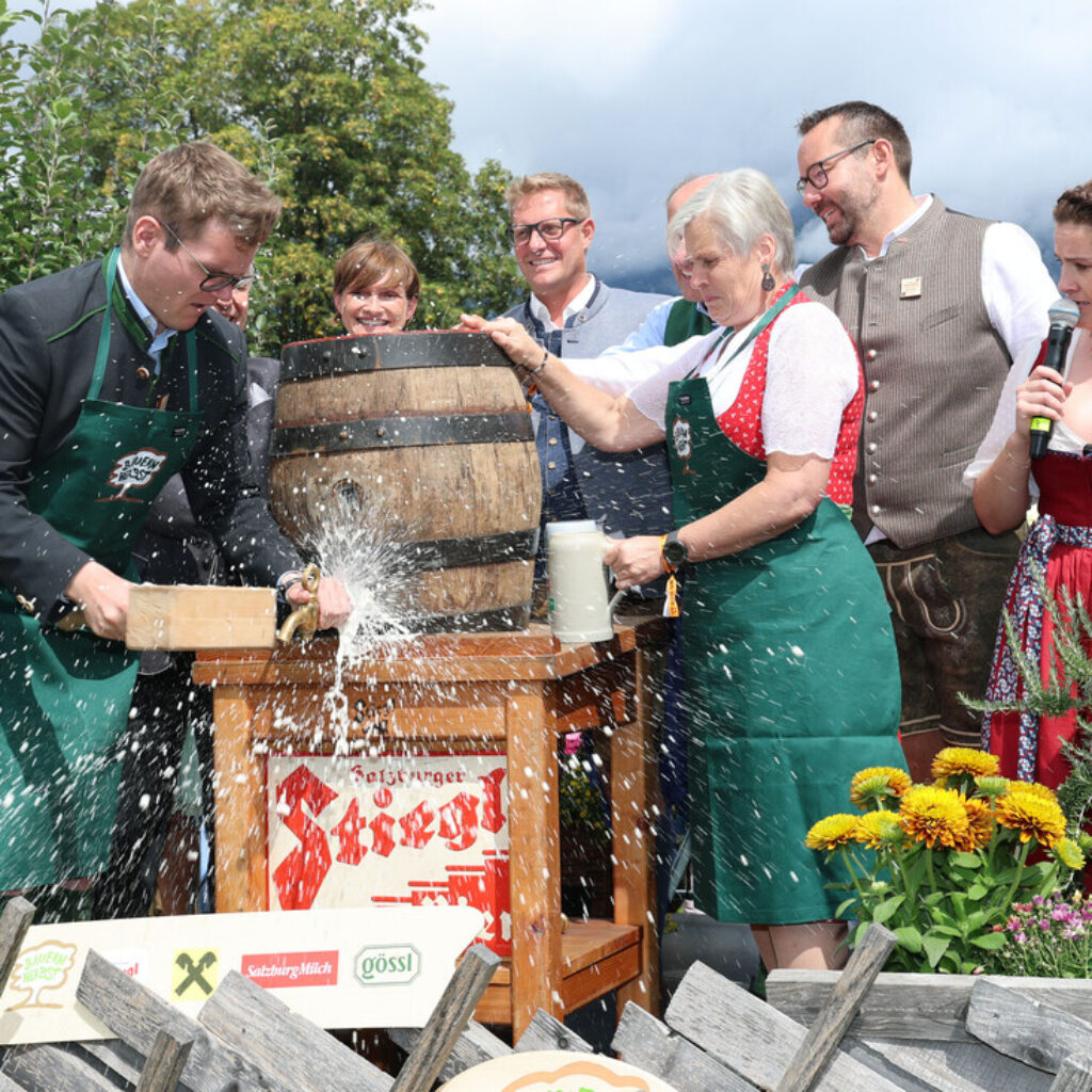 Ein Mann zapft ein Bierfass an, Schaum spritzt heraus, Menschen in traditioneller Kleidung lächeln und schauen zu, im Vordergrund sind Blumen zu sehen.