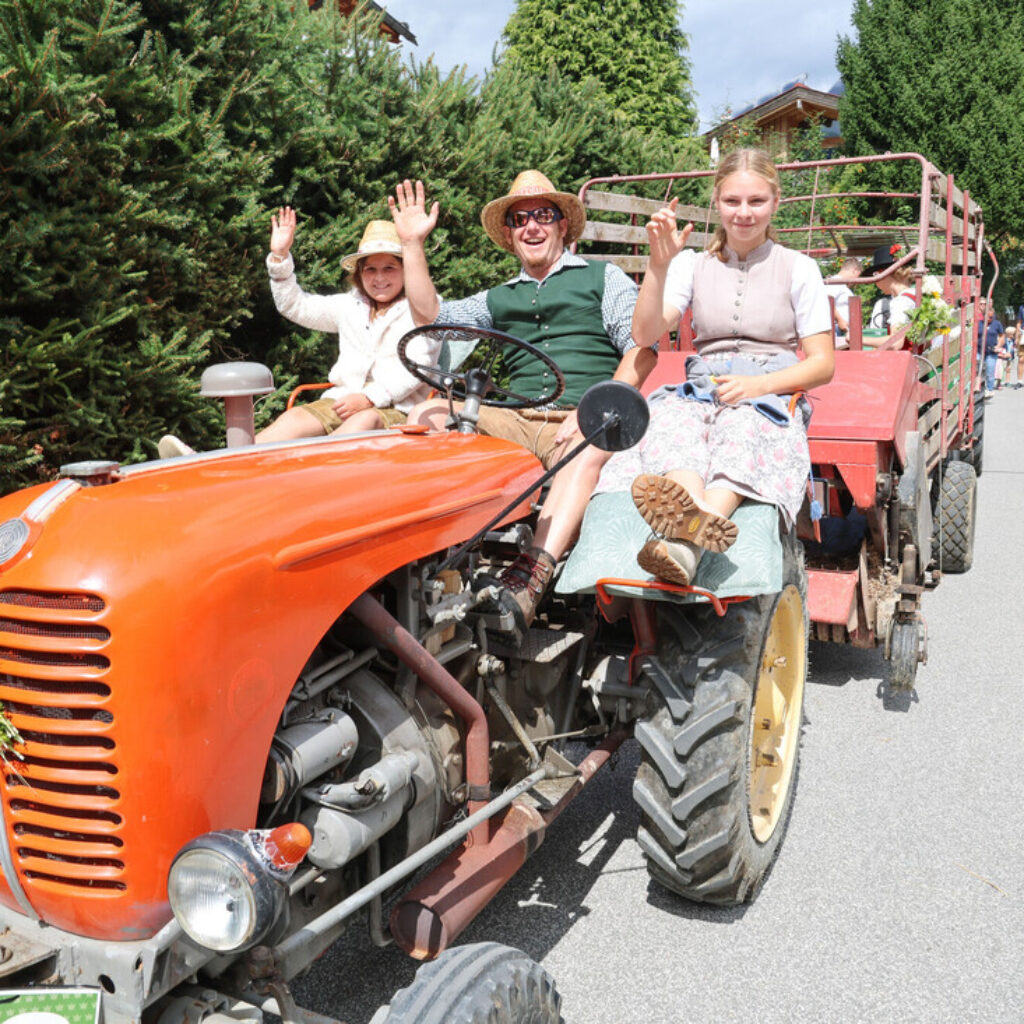 Drei Menschen in traditioneller Kleidung winken an einem sonnigen Tag während einer Parade von einem roten Traktor aus.