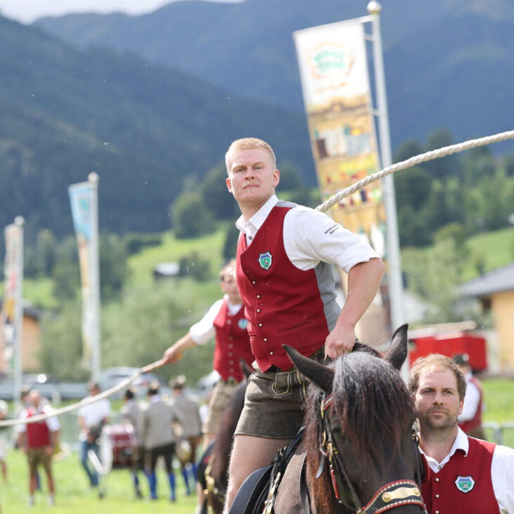 Männer in roten Westen reiten bei einer Freiluftveranstaltung in einer bergigen Gegend auf Pferden, im Hintergrund sind Transparente zu sehen.
