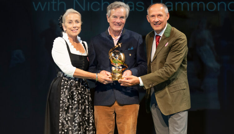 Drei Personen lächeln auf der Bühne, halten eine goldene Auszeichnung in den Händen, im Hintergrund ist ein „Sound of Music“-Schild zu sehen.