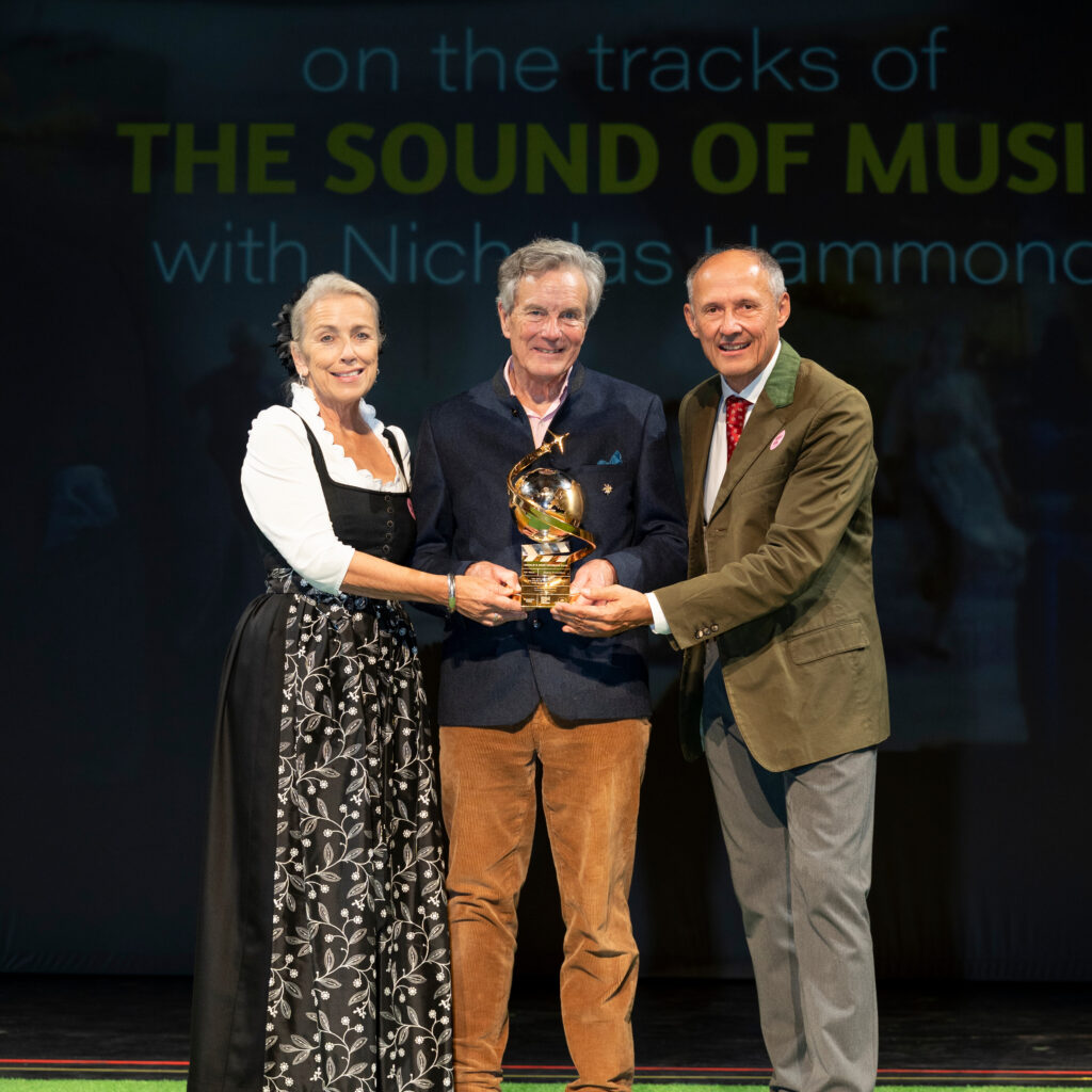 Drei Personen stehen auf der Bühne und halten eine goldene Auszeichnung in den Händen, hinter ihnen ist eine Kulisse aus „The Sound of Music“ zu sehen.