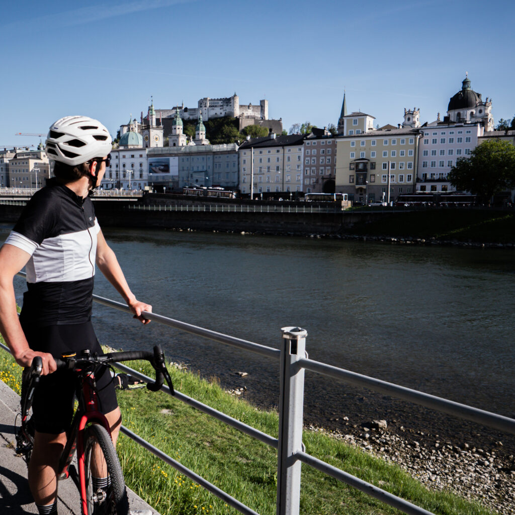 Ein Radfahrer genießt eine „Bike-Sommer“-Pause am Flussufer und bewundert vor dem Hintergrund des blauen Himmels eine Burg und historische Gebäude.
