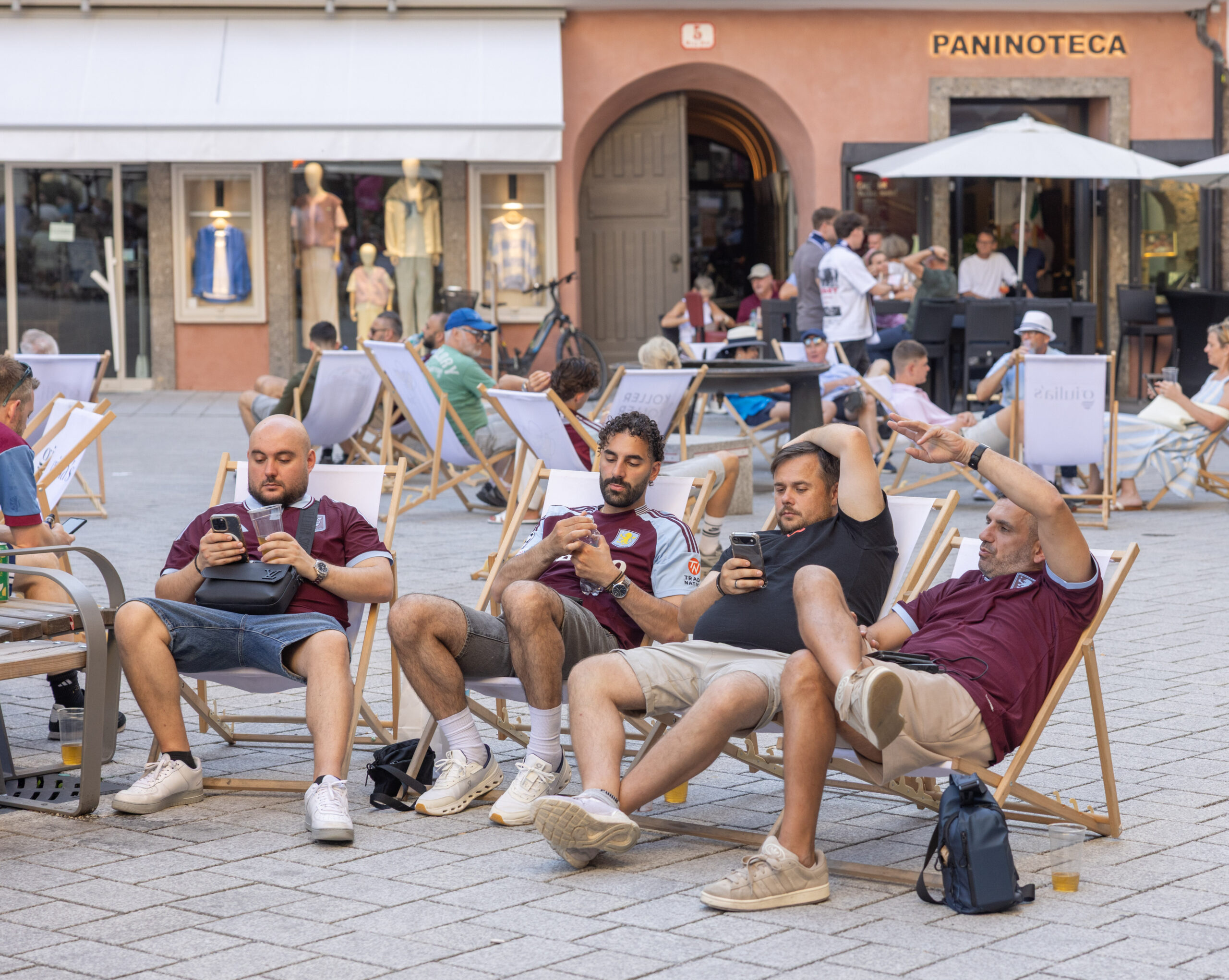 Vier Männer in kastanienbraunen Hemden entspannen sich draußen auf Liegestühlen und verfolgen jeweils auf ihren Handys die aktuellen Meldungen zum UEFA-Superpokal.