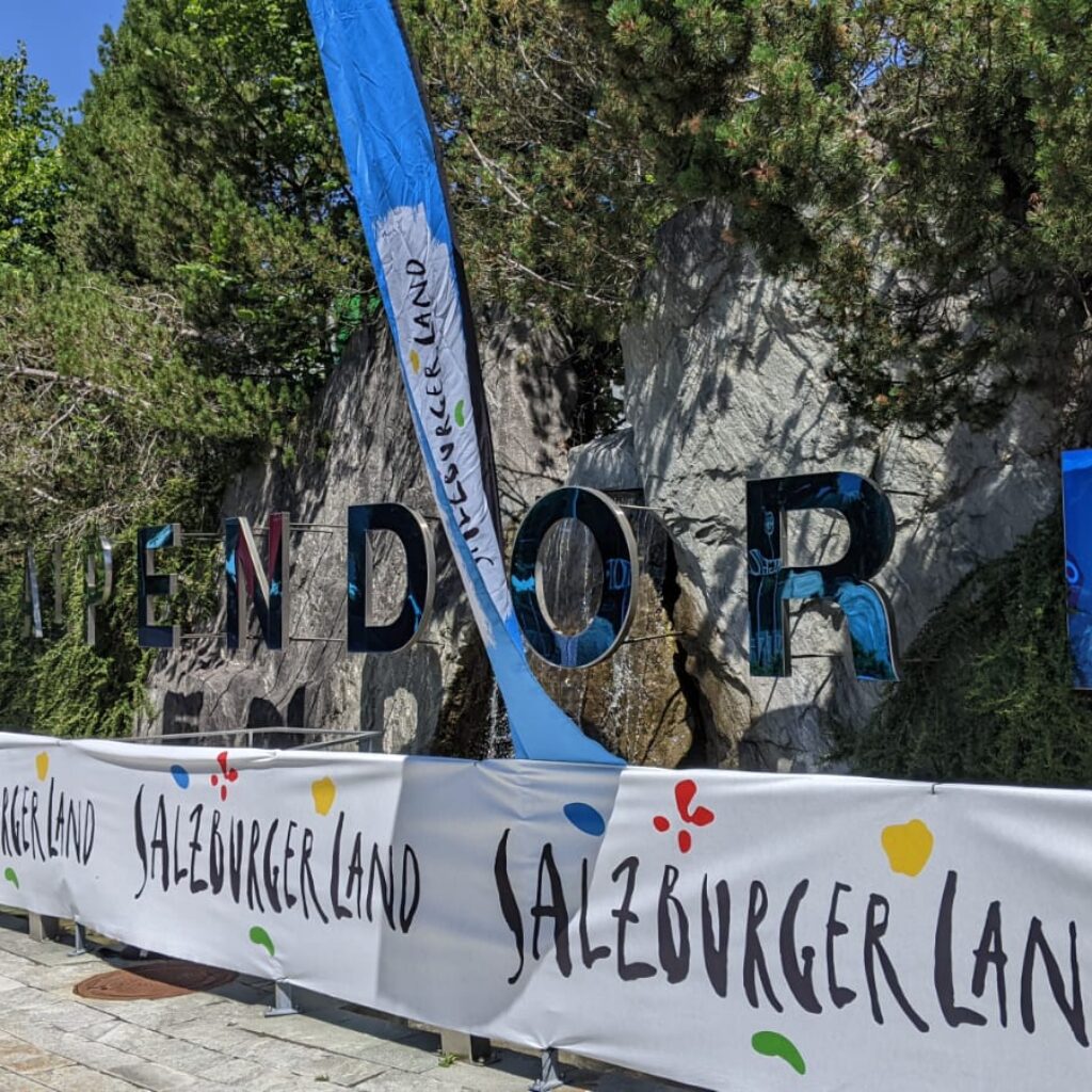 Großes Steinschild „ZELL AM SEE-KAPRUN“ mit SalzburgerLand-Bannern und „Bike-Sommer“-Fahnen an einem sonnigen Tag.