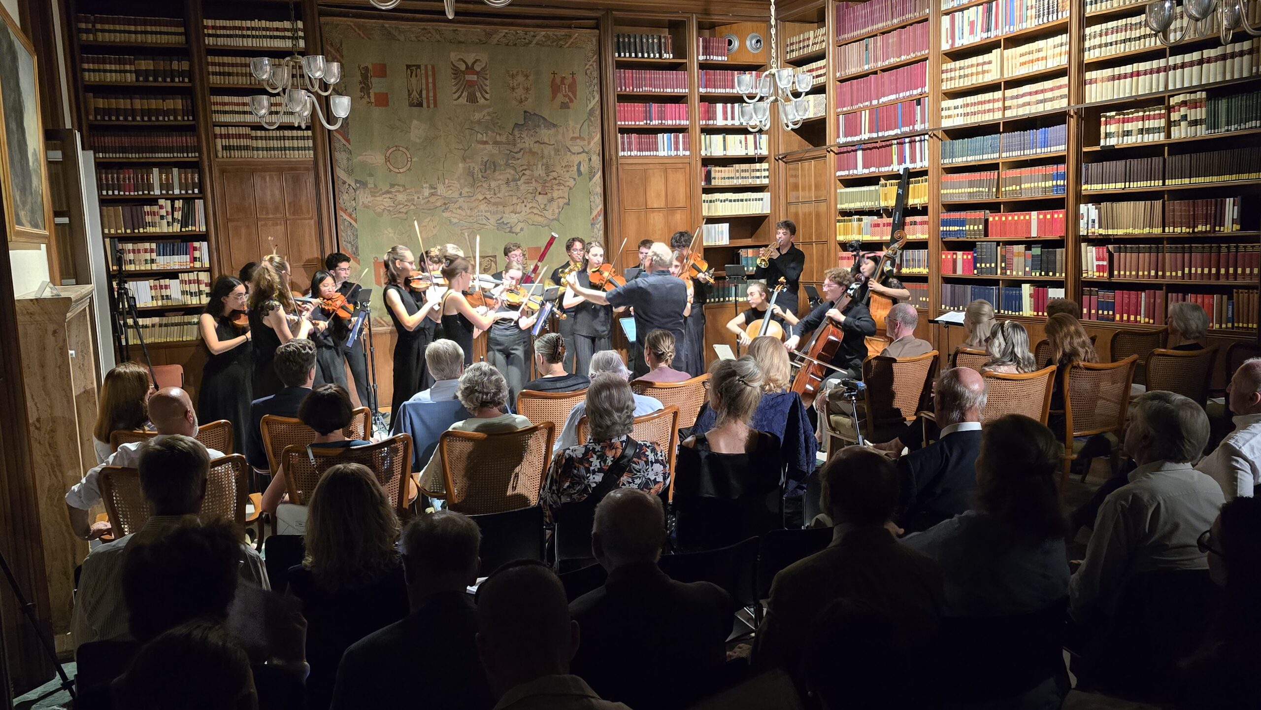 Ein Orchester spielt in einer Bibliothek vor Publikum, umgeben von hohen Bücherregalen und historischem Dekor.