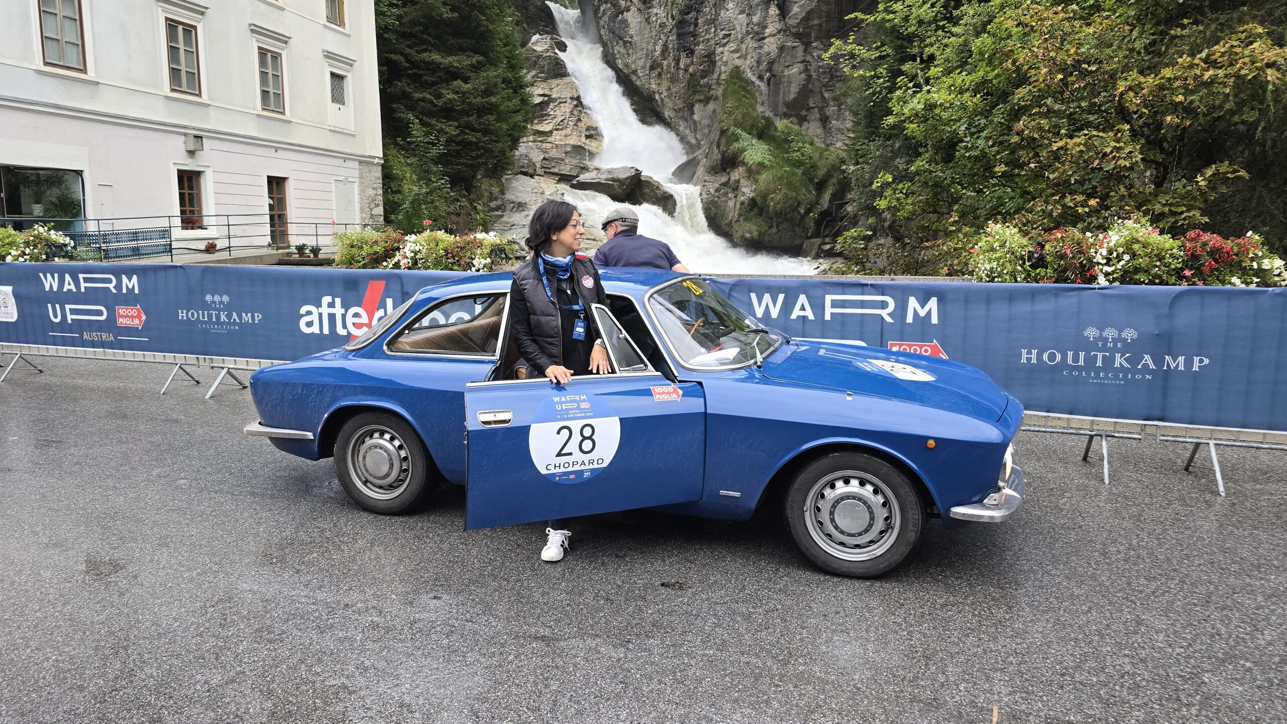Eine Person steht bei einer Oldtimer-Veranstaltung neben einem blauen Oldtimer mit der Nummer 28 in der Nähe eines Wasserfalls.
