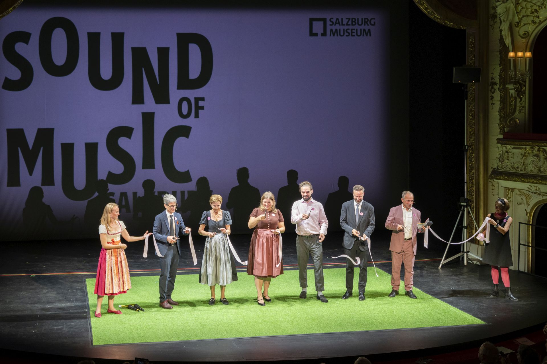 Sieben Personen auf der Bühne halten bei einer Feier vor der Kulisse des „Sound of Music“-Museums in Salzburg ein Band in den Händen.