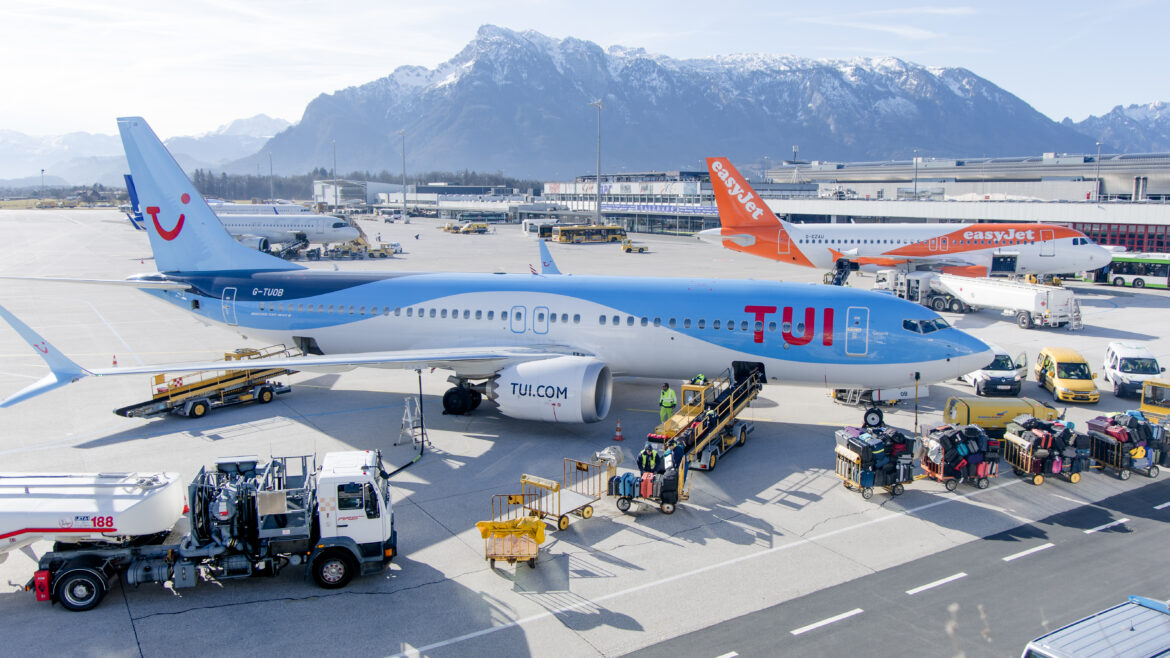 Ein TUI-Flugzeug auf dem Rollfeld des Flughafens, Gepäck wird verladen, im Hintergrund sind Berge und andere Flugzeuge zu sehen.