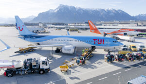 Ein TUI-Flugzeug auf dem Rollfeld des Flughafens, Gepäck wird verladen, im Hintergrund sind Berge und andere Flugzeuge zu sehen.
