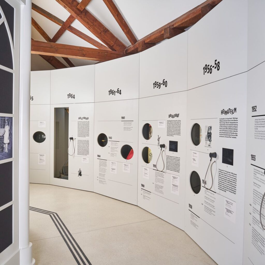 Eine geschwungene weiße Museumswand zeigt Exponate aus mehreren Jahrzehnten, Informationstafeln und Requisiten aus „The Sound of Music“; links ein Bogenfenster.