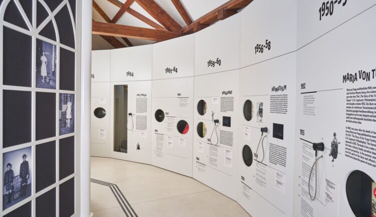 Eine geschwungene weiße Museumswand zeigt Exponate aus mehreren Jahrzehnten, Informationstafeln und Requisiten aus „The Sound of Music“; links ein Bogenfenster.
