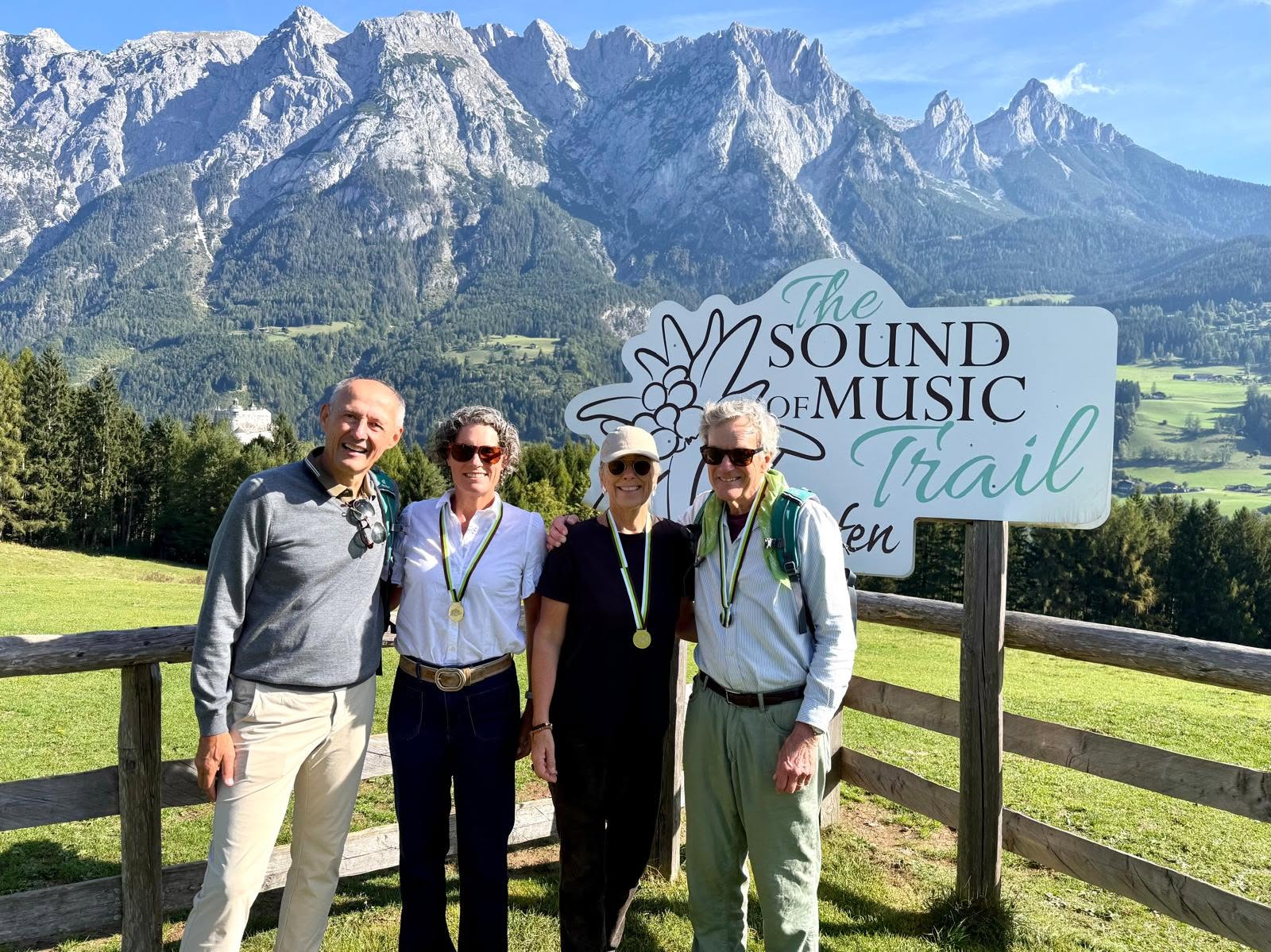 Vier Personen mit Medaillen stehen vor einem Schild des „Sound of Music“-Museumsrundgangs in Salzburg; hinter ihnen sind Berge und Bäume zu sehen.