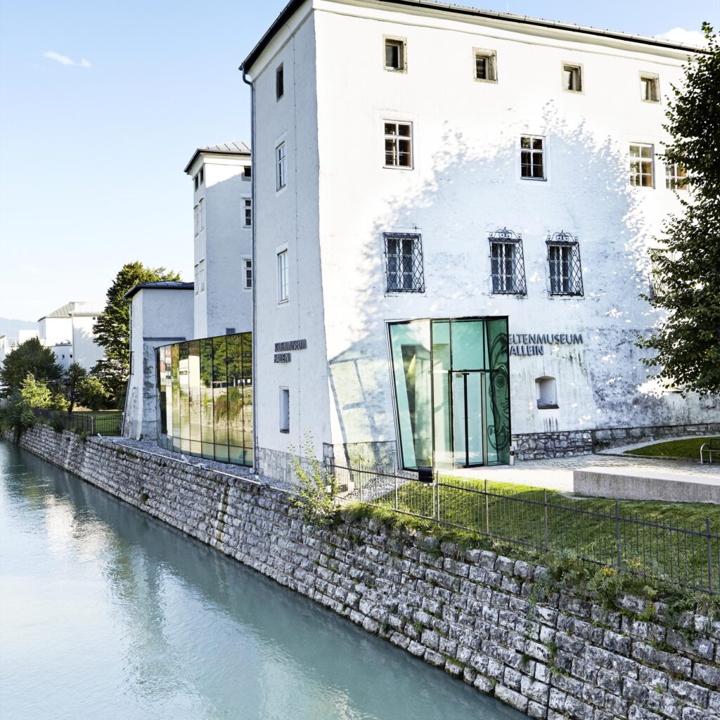 Moderner Glaseingang des „Sound of Music“-Museums in Salzburg neben einem ruhigen Wasserlauf unter strahlend blauem Himmel.