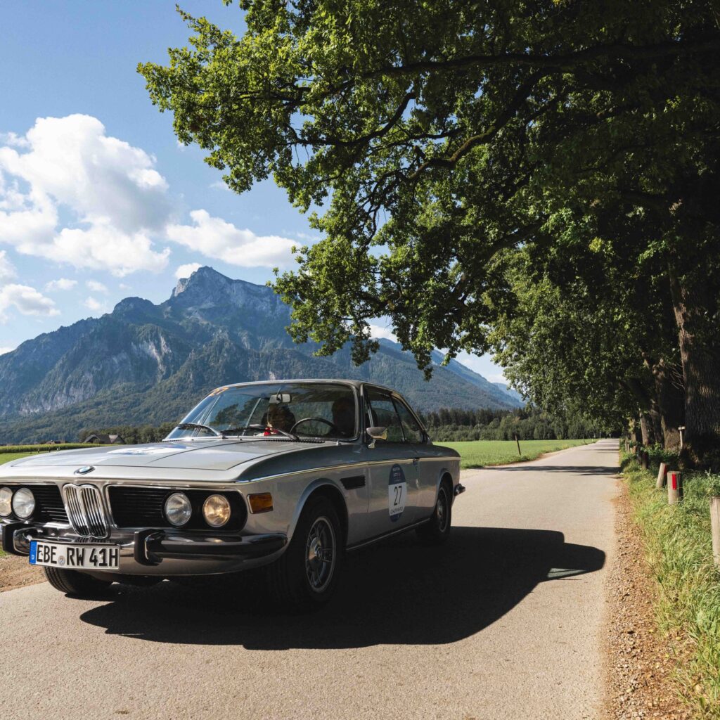 Ein klassischer silberner BMW fährt auf einer von Bäumen gesäumten Landstraße, im Hintergrund sind Berge und ein blauer Himmel zu sehen.