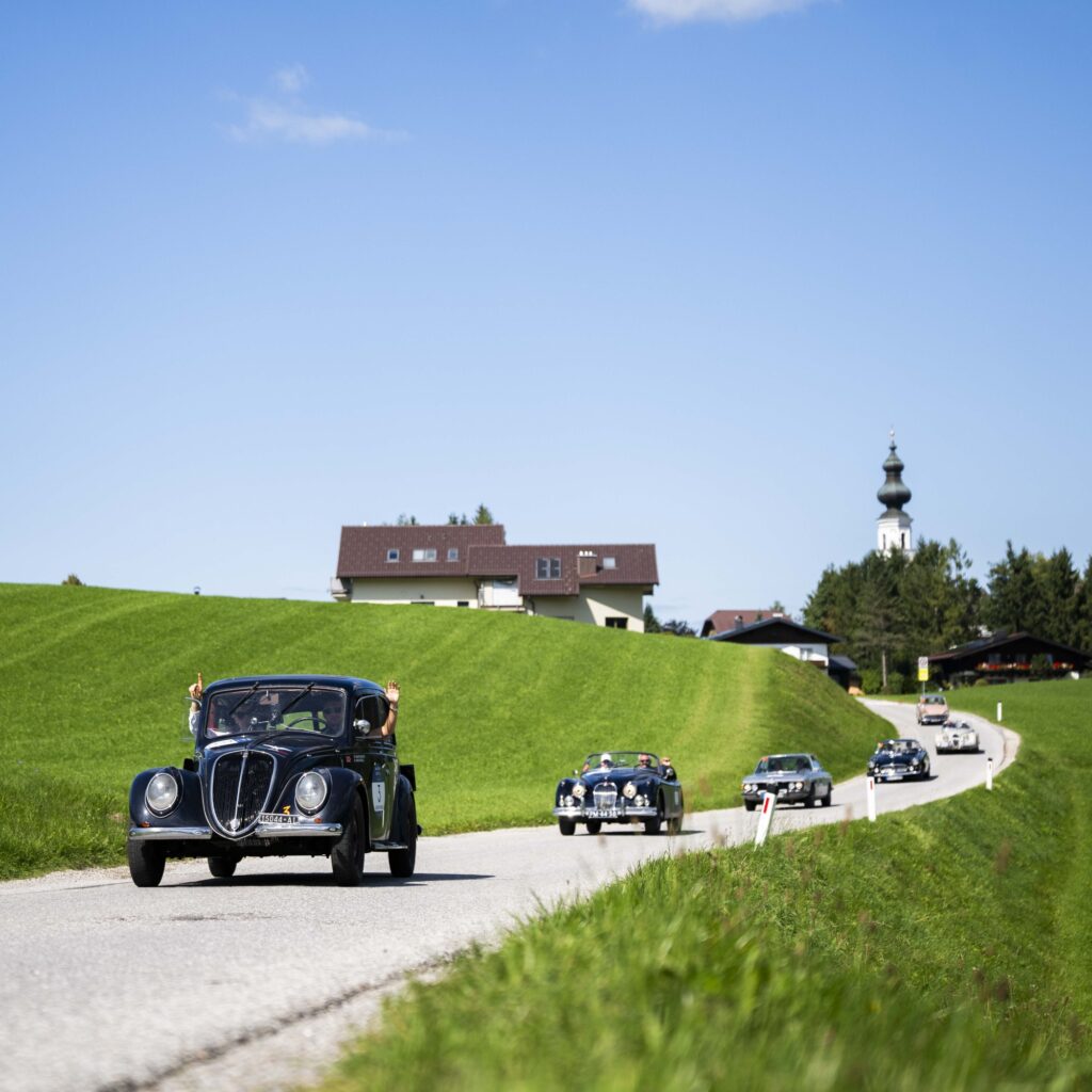 Oldtimer fahren auf einer Landstraße, im Hintergrund sind grüne Felder, Häuser und ein Kirchturm zu sehen.