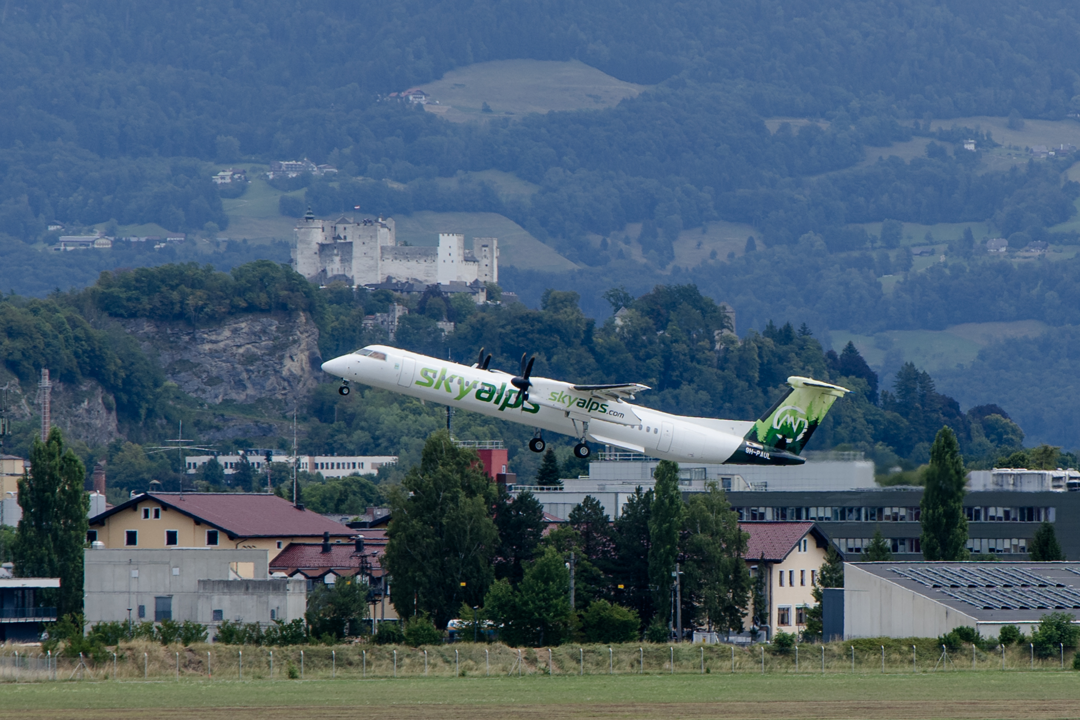 Ein Flugzeug von SkyAlps hebt in der Nähe von Gebäuden, Hügeln und einer Burg ab und macht damit auf den neuen Winterflugplan aufmerksam.