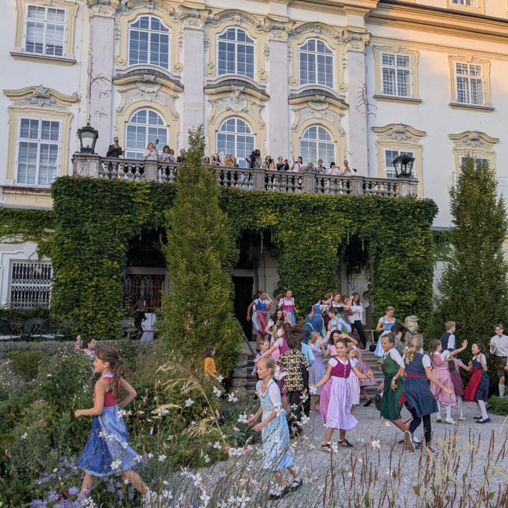 Kinder in bunten Kleidern laufen unter den Blicken der Zuschauer am mit Efeu bewachsenen „Sound of Music“-Museum in Salzburg vorbei.