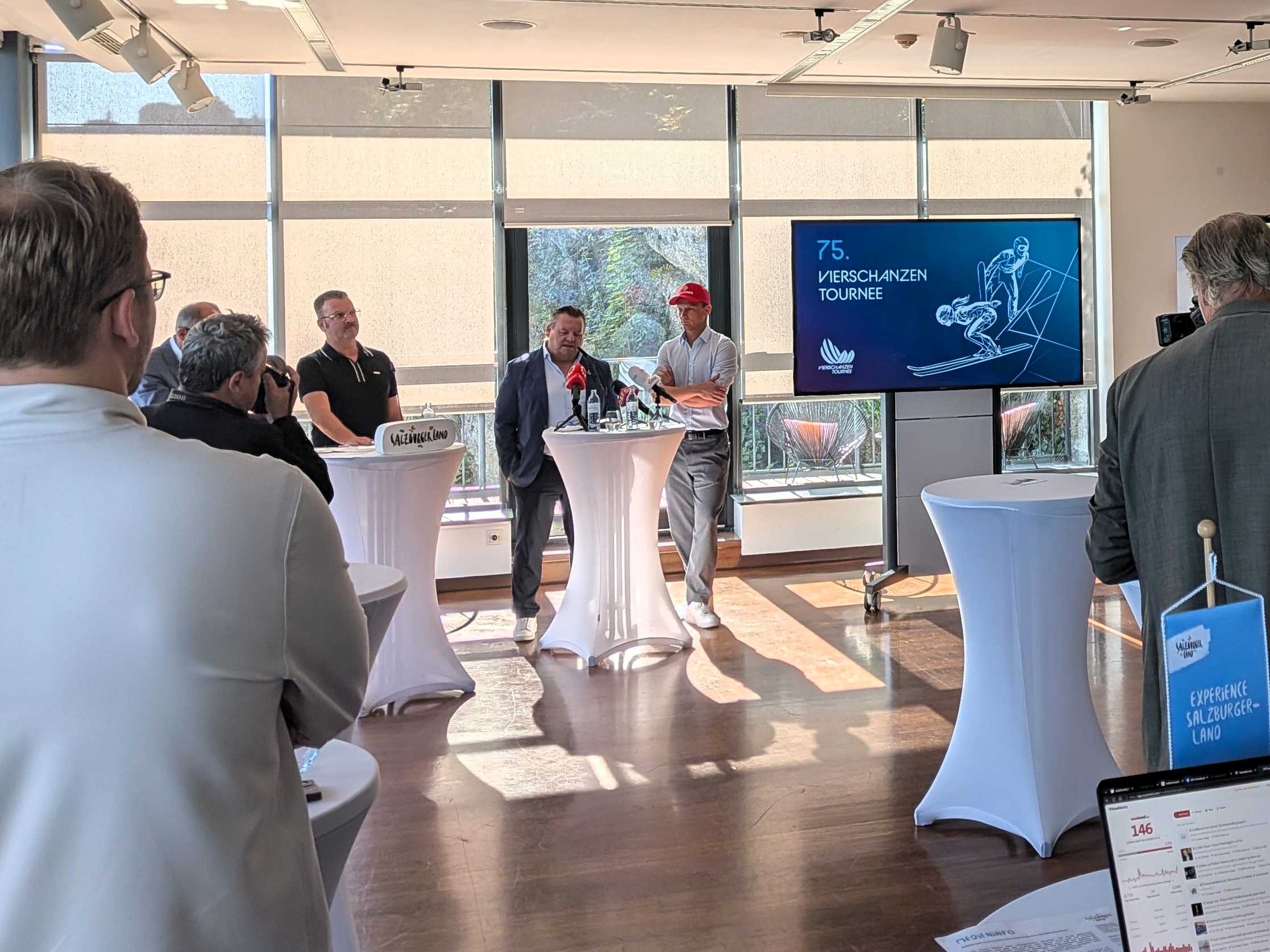 Bei einer Pressekonferenz zum 4-Schanzen-Turnier zum 75. Mal stehen zwei Männer hinter einem hohen Tisch.
