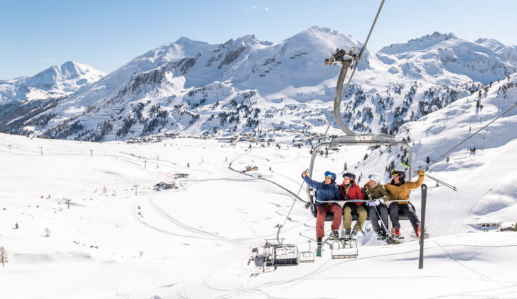 Vier Skifahrer sitzen auf einem Sessellift und machen ein Selfie vor der Kulisse der schneebedeckten österreichischen Berge.