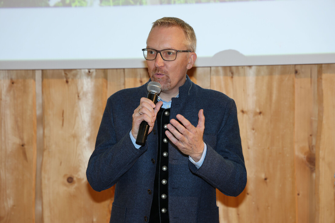 Ein Mann mit Brille spricht in ein Mikrofon, während er eine dunkle Lodenjacke trägt.