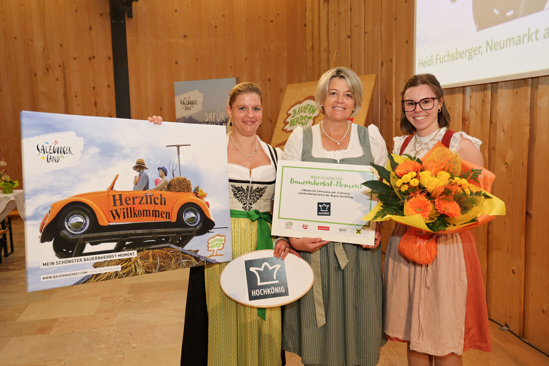 Drei Frauen in traditioneller österreichischer Tracht halten ein Werbeschild, einen Gutschein und einen Blumenstrauß in den Händen.