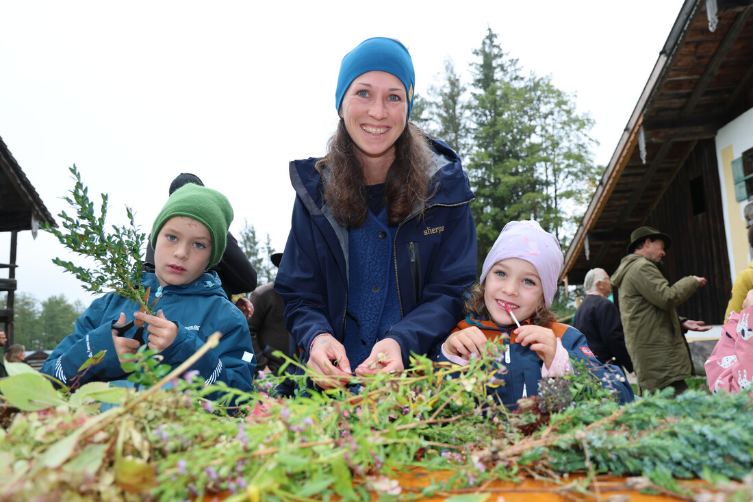 Eine Frau und zwei Kinder sitzen draußen an einem Holztisch und stellen Sträuße aus Wildblumen und Grünpflanzen zusammen.