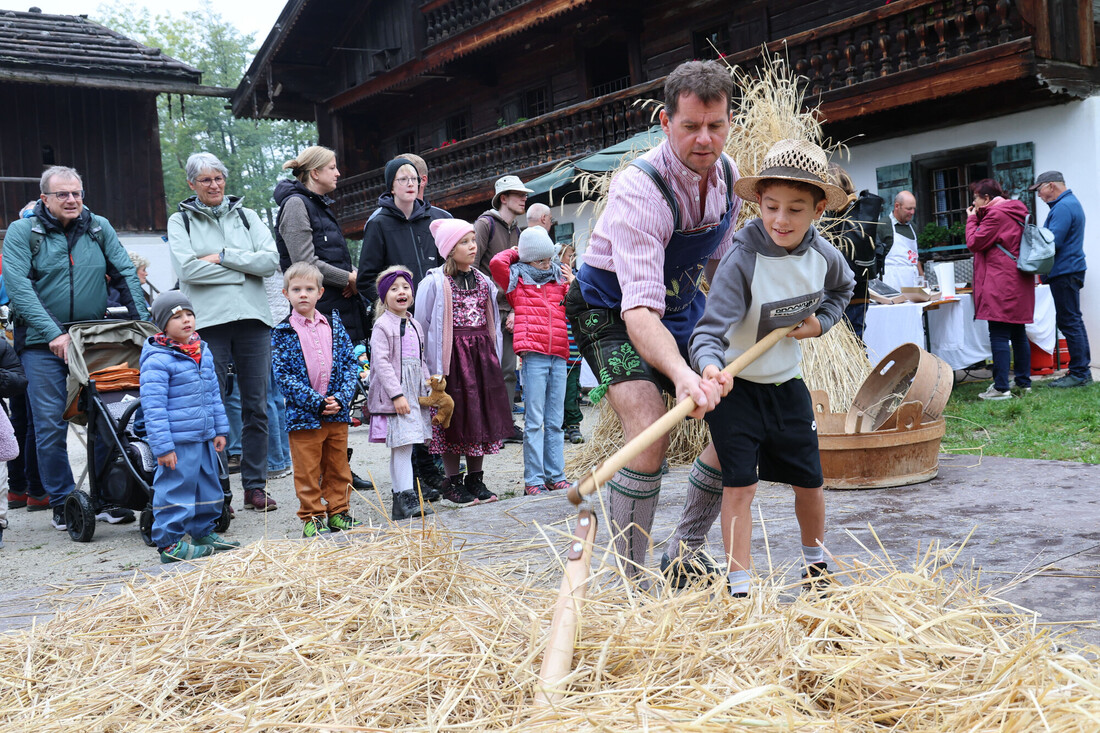 Ein Mann und ein Junge, die vor einem traditionellen österreichischen Holzhaus mit einer Sense Stroh mähen.