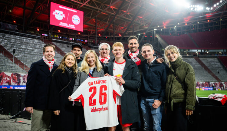 Eine Gruppe von Menschen steht auf einem Fußballplatz und hält ein weißes Trikot mit der Nummer 26 von RB Leipzig auf dem Rücken hoch.