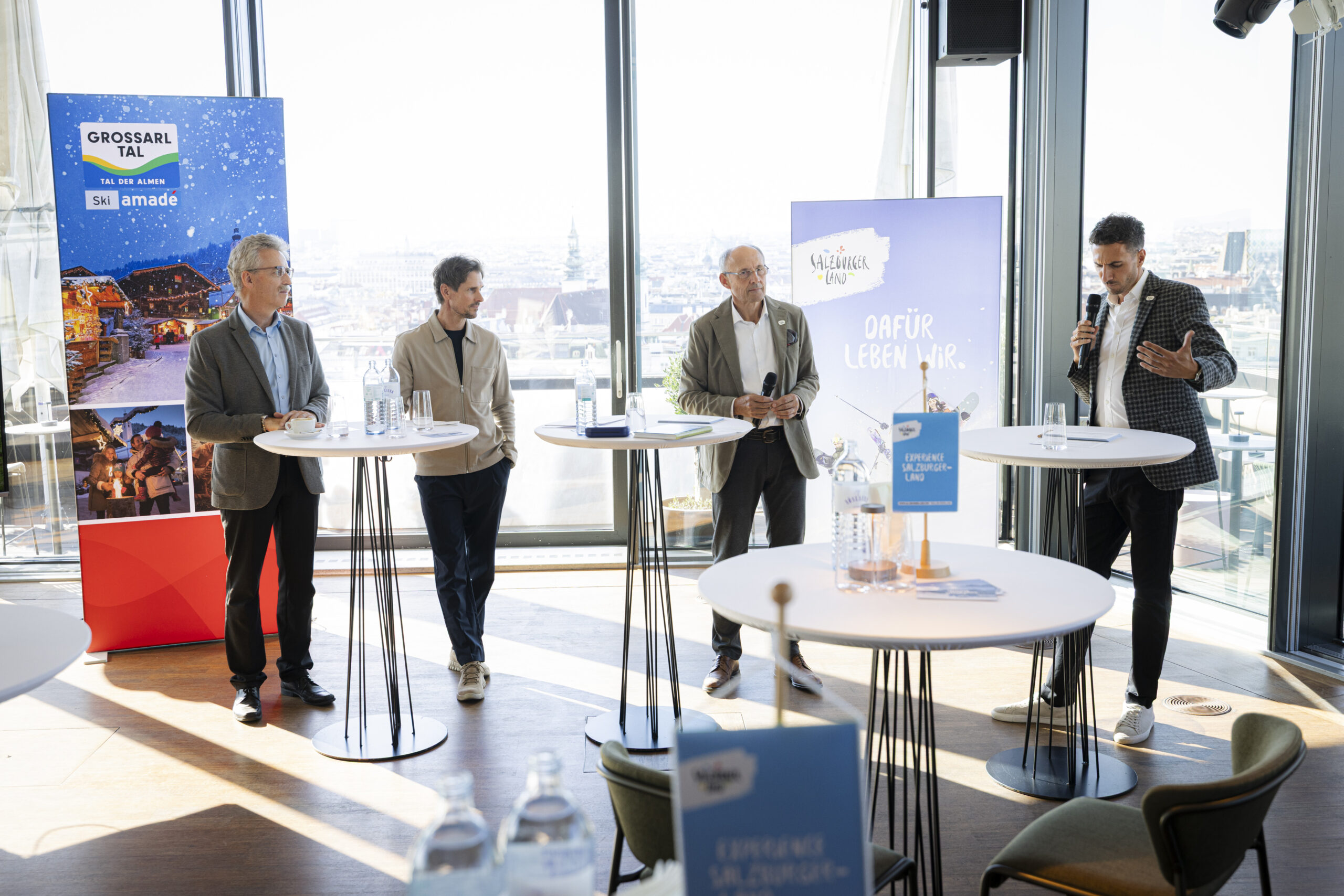 Vier Männer, die während einer Podiumsdiskussion an Stehtischen stehen, im Hintergrund eine Stadtlandschaft.