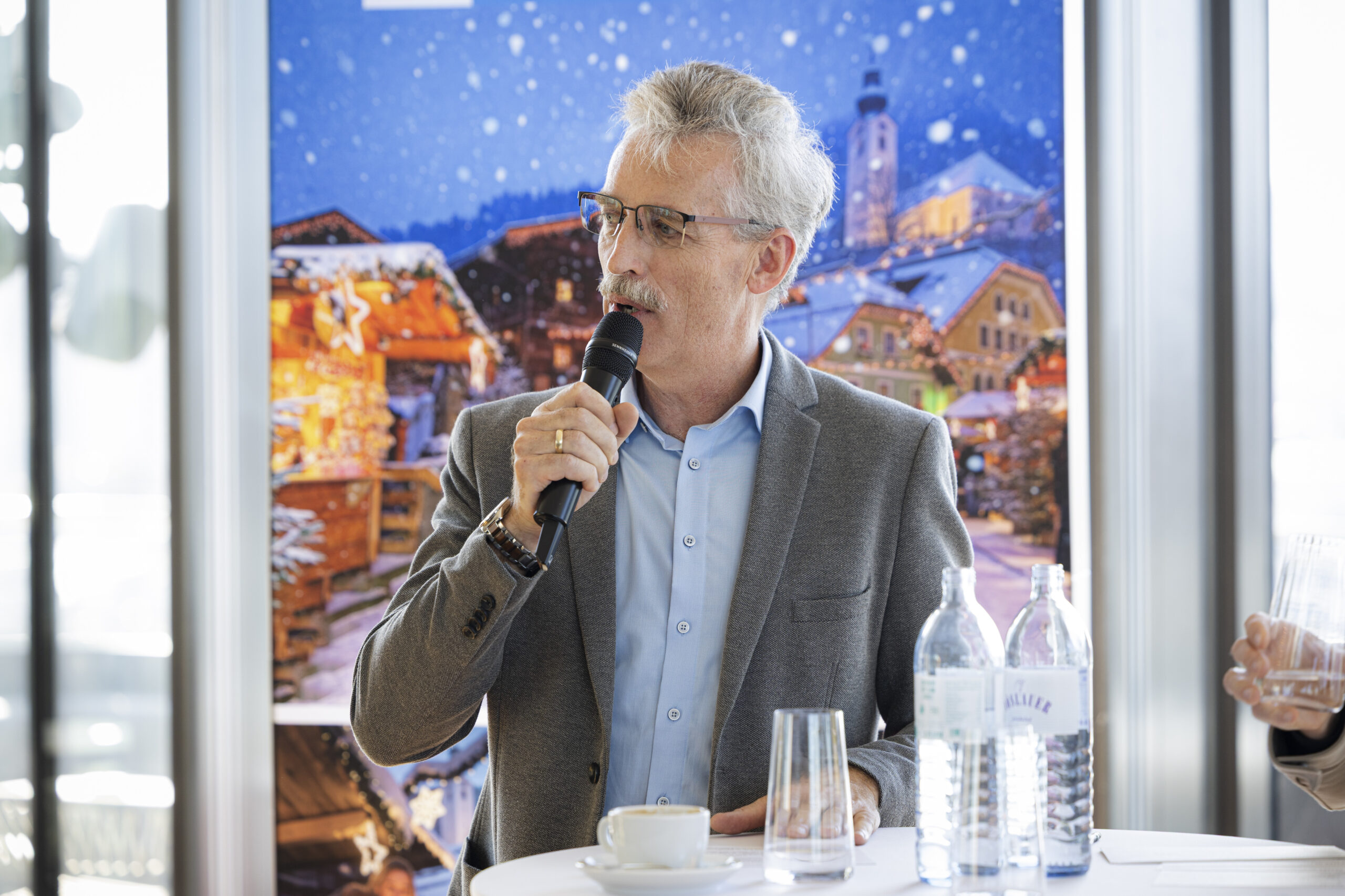 Ein Mann mit Schnurrbart und Brille spricht vor einer Kulisse, die einen Wintermarkt darstellt, in ein Mikrofon.