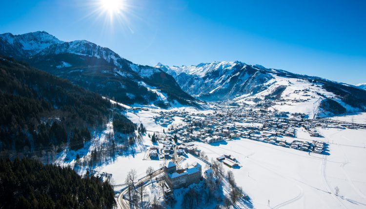 Ein sonniges, verschneites Alpendorf mit Bergen im Hintergrund und einem Wald im Vordergrund.